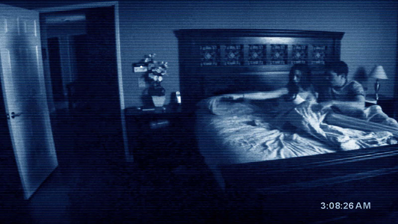 A tape recording of a couple sitting on a bed pointing at a large shadow on their bedroom doorway.