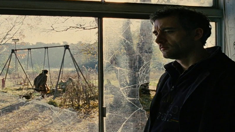 Clive Owen from 'Children of Men' standing next to a broken glass window and a swing set with someone sitting on it