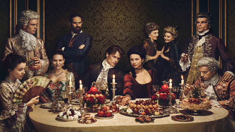 Many cast members from 'Outlander' on Starz in very extravagant clothing gathered around a table